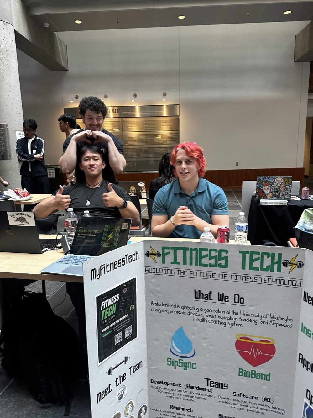 Three Fitness Tech members at the club table in front of the tri-fold poster, one making a heart over another's head